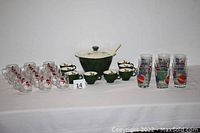 Full lot showing green hot cider bowl with lid and ladle, 8 matching holly mugs, 12 colorful holiday-themed glass tumblers, and 12 clear snowman glass mugs all arranged on table.