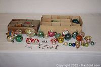 Wide shot showing the two boxes of vintage Christmas ball ornaments and many loose balls in various colors and patterns on a white surface