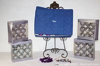 Four boxed sets of Sandra Lee Christmas tree balls in metallic and muted colors, arranged around a blue quilted case on a decorative stand, with some loose decorations visible at base