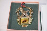 Close-up of 1996-1997 Official Federal Duck Stamp holiday ornament featuring a brass/gold-tone hinged wreath with bird and inserted duck stamp image, resting in original pink box with green velvet lining, with red hanging ribbon attached.