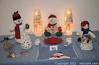 Wide view of all snowman decorations on blue cloth including vintage snowmen lights, plush dolls, snow globe, candle holders, pot holder and metal ornament.