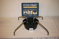 Front view of the Stand Strait tree stand with four metal legs, green water reservoir, and label number 114 arranged on white surface