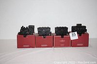 Four cast iron black train-themed stocking holders displayed on their original red boxes in a single row.