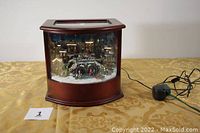 Front view of wooden display case with Victorian village diorama illuminated inside, showing power cord attached