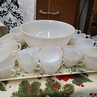 Milk glass punch bowl set with punch bowl, stand, and many matching milk glass cups arranged on a tray with holiday-themed linens in the background.
