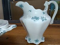Front and side view showing the pitcher with white porcelain and green floral designs, decorative spout, and shaped base.