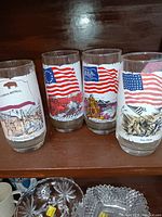 Photo showing set of 4 Coca-Cola branded collectible glasses with American flags and California Republic flag along with battle scenes.