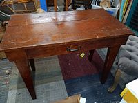 Front and angled view of the primitive wooden desk showing its reddish-brown surface, metal handle on the drawer, and sturdy legs.