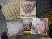 Five vinyl records stacked on carpet and rug. Albums by Rod Stewart and Olivia Newton-John visible.