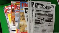 Photo showing a stacked array of 15 vintage car magazines, primarily featuring Auto Restorer issues with black and white covers and some colored Super Chevy and other automotive magazines behind them.