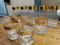 Set of vintage barware glasses arranged on wooden surface, showing five lowball glasses with wide gold rims in front and five taller highball glasses with similar gold rims behind.
