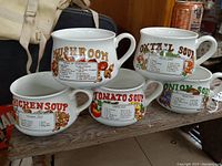 Five vintage ceramic soup cups arranged on wooden shelf, each labeled with a soup recipe and colorful illustrations. Cups include onion, oxtail, mushroom, chicken, and tomato soups.