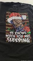Photo showing the front of a black XL T-shirt with a graphic of Freddy Krueger wearing a Santa hat and the text 'He knows when you are sleeping'.