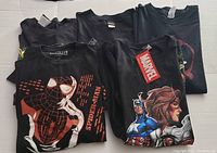 Lot of 5 black Marvel graphic T-shirts folded and stacked, showing front Marvel character designs including Spider-Man, Captain America, and Black Widow. Shows tags on one shirt.