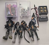 Four KISS band member action figures laid out, including one holding dragon-shaped staff, guitars, and speaker amplifier accessories in small boxes
