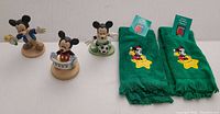 Three Mickey Mouse figures in different outfits and two green holiday-themed Mickey towels with tags arranged on white background.