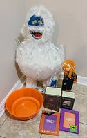 Photo of white furry yeti plush figure standing on the floor, small scarecrow doll, orange plastic bowl, and four Halloween-themed books on the floor against wall