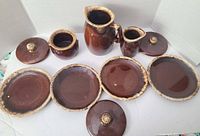 Overview of 9 items including 2 pitchers, 3 lids, and plates arranged on white background showing brown drip glaze with some wear and chips.