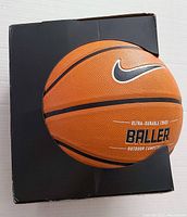Front view of the Nike Baller basketball on top of its original black box, showing the orange surface with black lines and the Nike swoosh, with text stating 'Baller Outdoor Competition'.