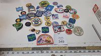 Photo showing an assortment of camping and scouting badges of various shapes and colors spread out on a white surface with ruler for scale.
