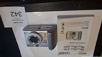 Silver Canon PowerShot A70 digital camera with zoom lens and control dial, displayed with the user manual and a lot card.