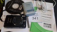 Canon PowerShot ELPH 510 HS camera placed on user manual, software CD, and battery charger beside it with a lot number card.