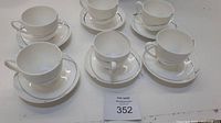 Top view of 6 white cups each placed on a matching white saucer, all arranged in two rows on a white surface with lot tag.