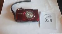 Front view of red Nikon Coolpix L26 showing lens, branding and wrist strap, on a white surface with lot number card.