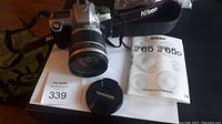 Nikon F65 film camera with attached Tamron lens, camera strap, lens cap, instruction manual, and a B+W 82E 010 1K lens filter for scale.
