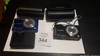 Two Panasonic Lumix cameras, black and blue, with battery charger and batteries displayed on a table.