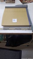 Photo showing the full paper cutter on a table, displaying the beige cutting board with grid lines and the gray cutting arm with blue handle.