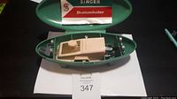 View of Singer Buttonholer device inside open olive green plastic case with instruction leaflet visible at the top.