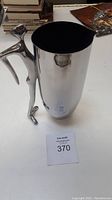 Stainless steel pitcher with sculptural human figure handle against a background of books and artwork.