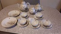 Wide shot showing the full tea set including teapot, creamer, sugar, cups, saucers, and plates arranged on a table with fabric covering.