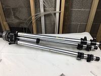 Full view of folded aluminum tripod with black clamps and rubber feet on legs, placed on white surface with background showing basement wall.