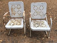 Front view of two vintage metal outdoor chairs side by side on dirt ground with pine needles, showing white painted finish and decorative cut-out pattern on backrest and seats, displaying rust and weathering.