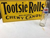 Yellow tin Tootsie Rolls sign with black text alongside Pepsi-Cola glass bottle on white surface