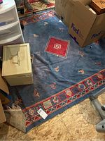 Photo showing a blue rug with red square and geometric patterns, visible dirt and wear, under storage items.