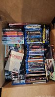 Box containing a mix of DVDs and Blu-ray discs including TV series and movie titles piled inside a cardboard box.