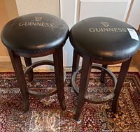 Pair of Guinness bar stools with wooden legs and black cushioned seats featuring Guinness logo; some scuffing on seats visible.