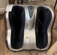 Front angled view of the Junior King Kong foot massager showing dual foot compartments with fabric lining and zippers, silver casing, and power cord.