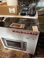 KitchenAid compact oven in original packaging with power and size details visible, next to French press and mini food chopper on table.