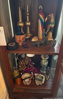 Photo of various figurines and sculptures arranged on two wooden shelves showing Royal Doulton Toby mugs, brass finials, carved wooden birds, small turtle figurines, knight figurine, and other decorative items.