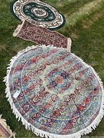 Three rugs displayed outdoors on grass: one large round rug with multicolor floral medallion design and cream fringe, smaller green and cream round rug, and red/blue rectangular runner rug.