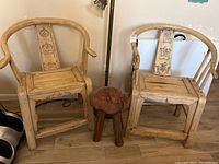 Pair of rustic arm chairs flanking a live edge wood stool. Chairs have carved backs and curved arms; stool has natural wood edge top and three legs.