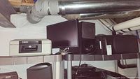 Top shelf containing a Samsung computer monitor, Sony SA woofer speaker, and Sentry safe lock box all placed on a metal shelf under visible pipes in basement.