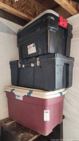 Three stacked storage containers: maroon bin with white lid at bottom, black footlocker in the middle, and black bin with red latch on top.