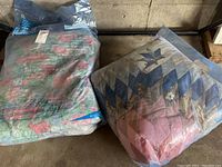 Two vintage quilts in sealed clear plastic bags on a concrete floor, one with a floral pattern and the other with patchwork star design.