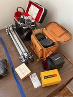 Photo shows two camera cases, a silver metal tripod, and camera accessories arranged on a wooden surface.