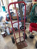 Full view of red metal hand dolly showing frame, wheels, and base plate, with surrounding garage items